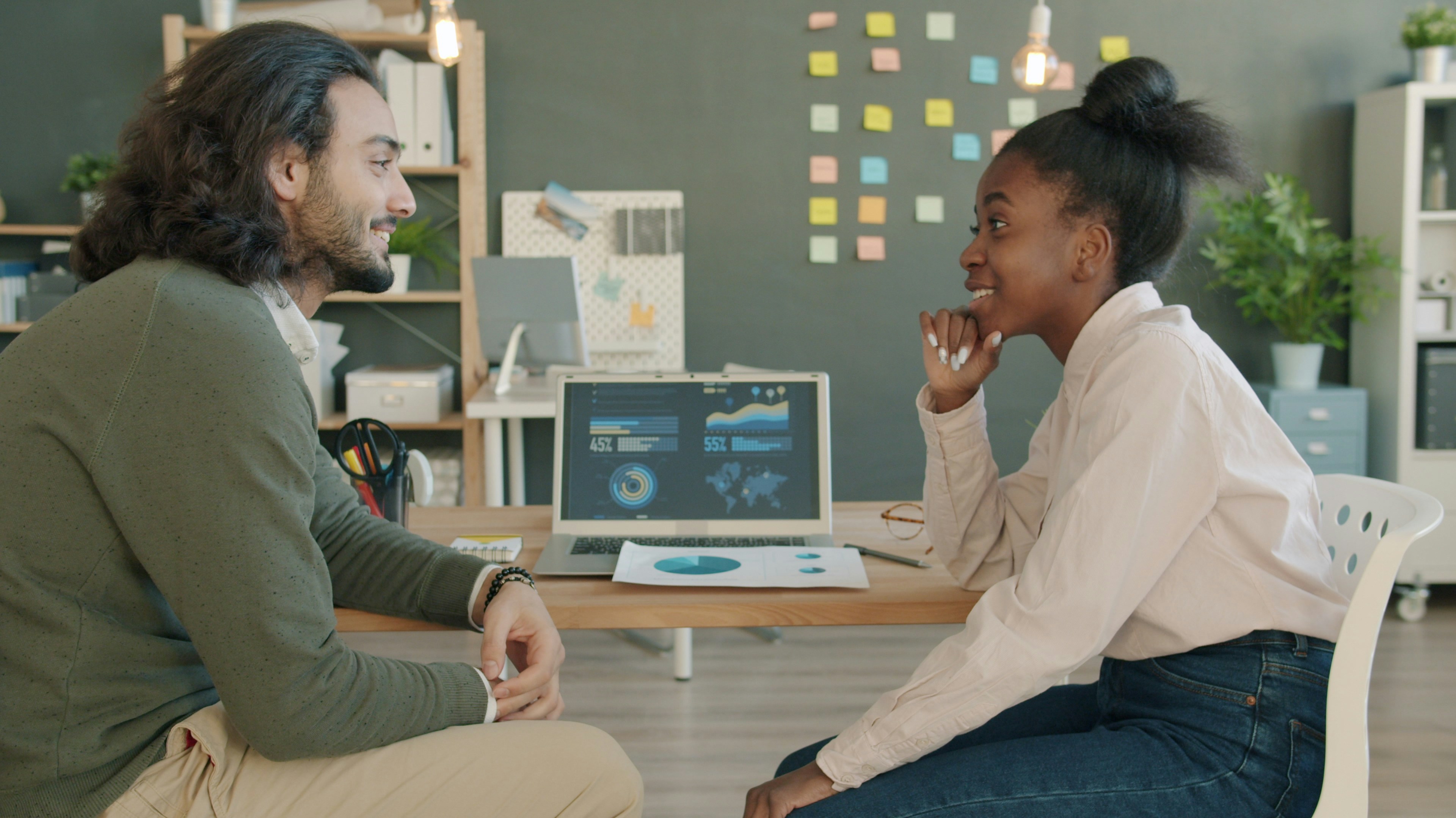 Arab guy and Afro-American girl are chatting and laughing looking at laptop screen in office. Modern technology, teamwork and workplace concept.