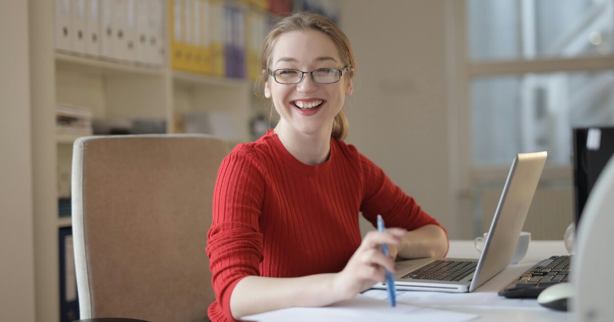 In-Blog-Thumbnails-41 Young student woman on her laptop smiling at the camera, wearing a red sweater.